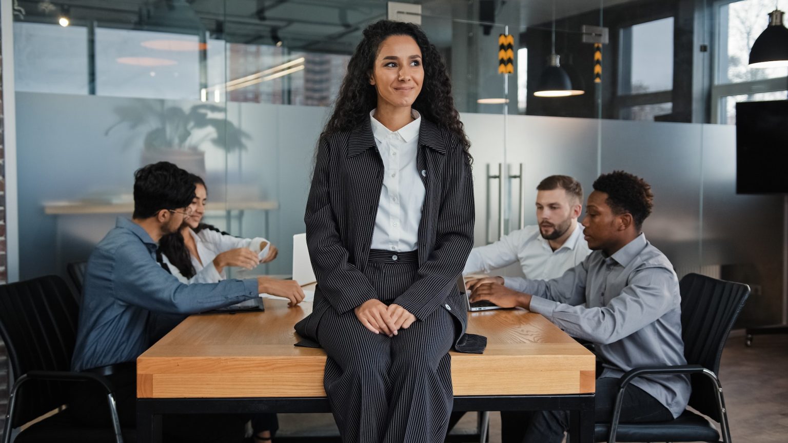 Pensive smiling businesswoman female leader boss woman sitting on table in office thinking